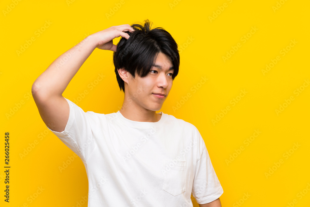 Asian man over isolated yellow wall having doubts while scratching head Asian man over isolated yellow wall having doubts while scratching head