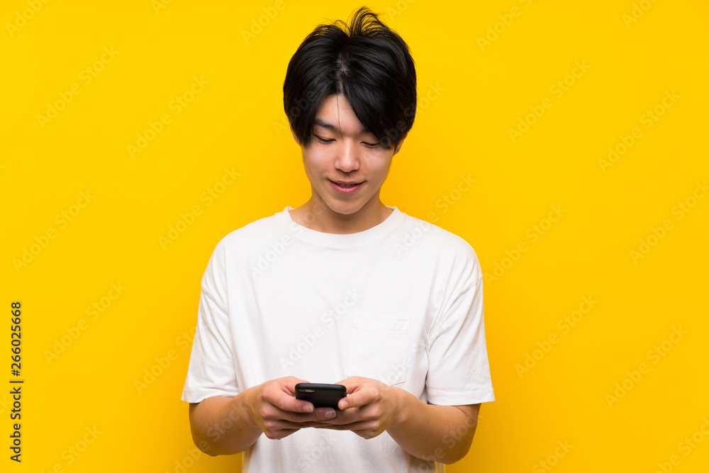 Asian man over isolated yellow wall sending a message with the mobile Asian man over isolated yellow wall sending a message with the mobile