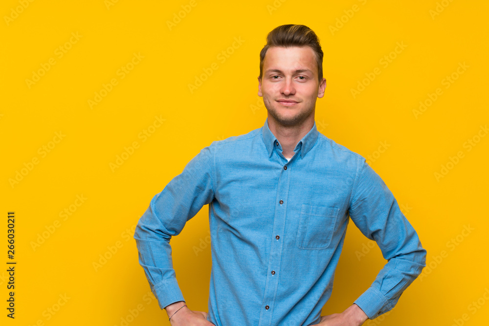 Blonde man over isolated yellow wall posing with arms at hip and smiling Blonde man over isolated yellow wall posing with arms at hip and smiling