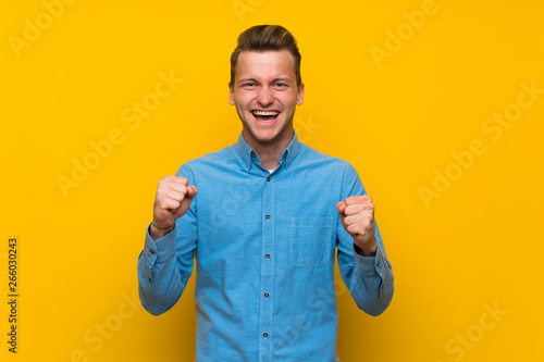 Blonde man over isolated yellow wall celebrating a victory in winner position