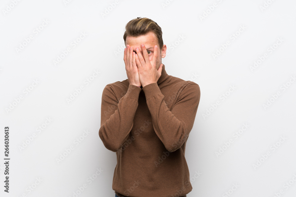 Blonde man over isolated white wall covering eyes by hands and looking through the fingers Blonde man over isolated white wall covering eyes by hands and looking through the fingers
