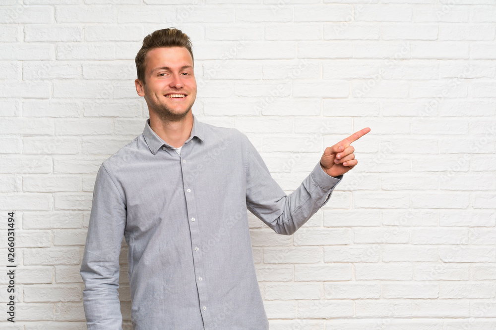 Blonde man over white brick wall pointing finger to the side Blonde man over white brick wall pointing finger to the side