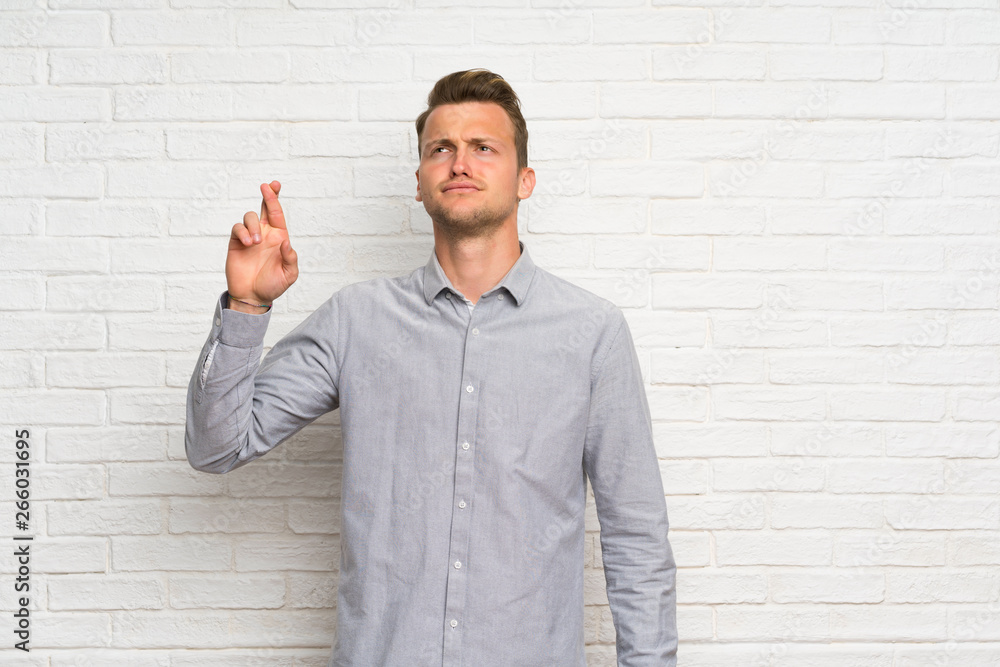 Blonde man over white brick wall with fingers crossing and wishing the best Blonde man over white brick wall with fingers crossing and wishing the best