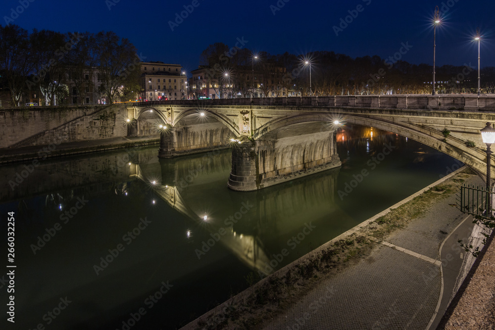 Ponte Sisto or Aurelius bridge is a stone road bridge in the historic center of Rome over the river Tiber at night with lighting. Path on the riverbank with lateral view