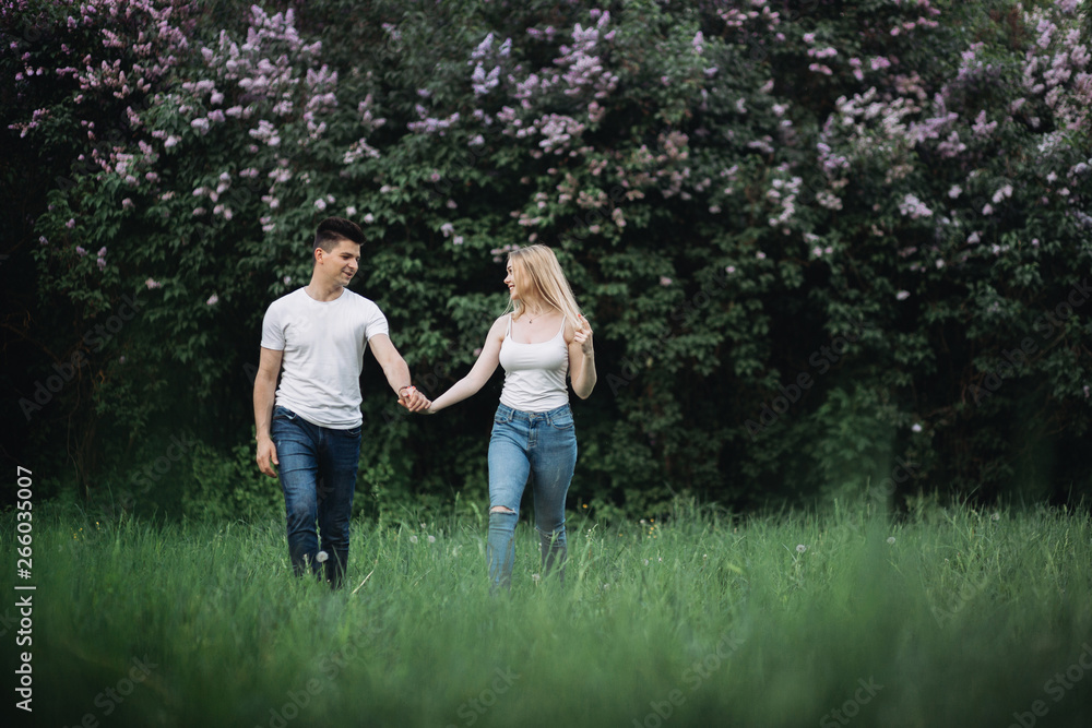 Fototapeta premium A young couple in love holding hands and walking forward in front of a flowering bush