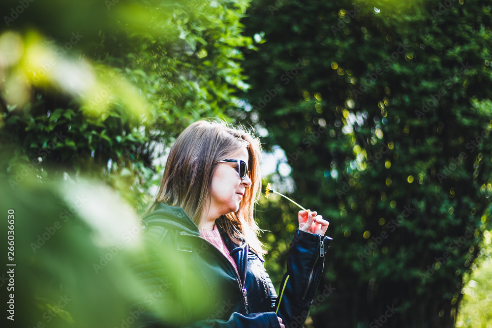 Young woman with sunglasses and a leather jacket smelling a small dandelion – Girl holding a little yellow flower on a bright spring day with trees in the background Young woman with sunglasses and a leather jacket smelling a small dandelion – Girl holding a little yellow flower on a bright spring day with trees in the background