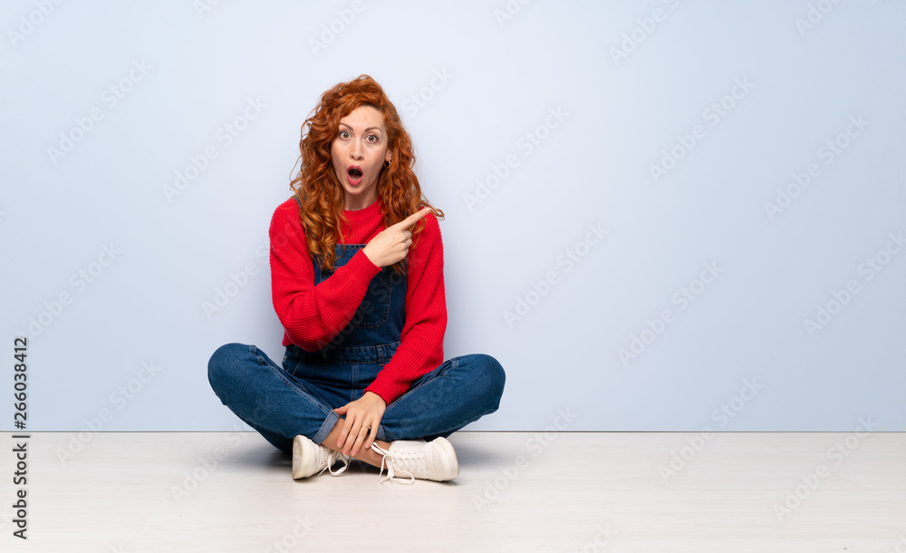 Redhead woman with overalls sitting on the floor surprised and pointing side Redhead woman with overalls sitting on the floor surprised and pointing side