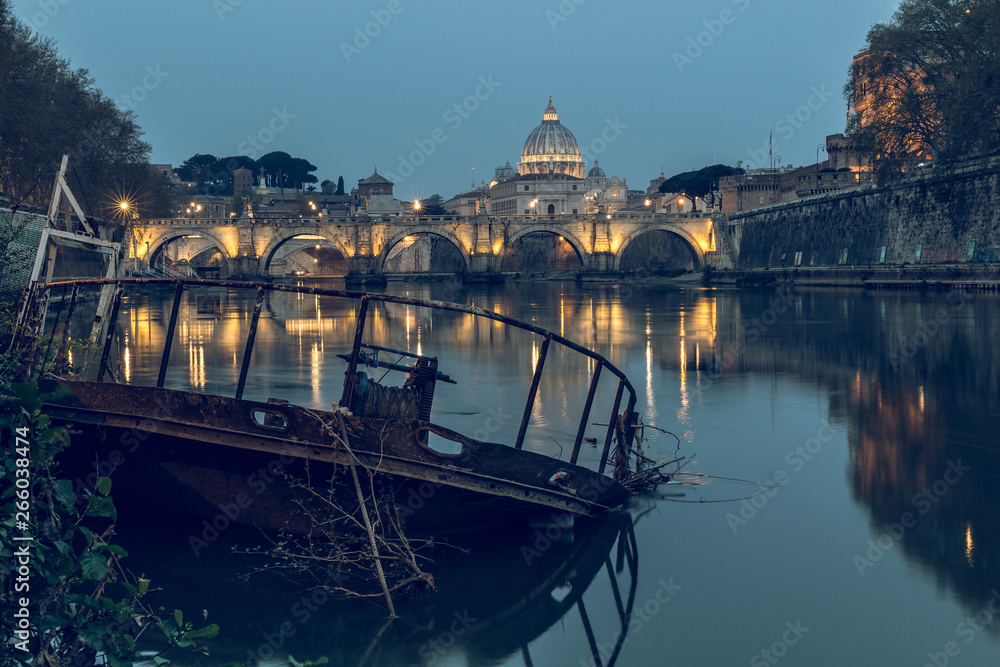 River Tiber in Rome and St Peters Cathedral at night. In the foreground a sunken ship at the pier. Buildings are illuminated. Shore with trees and reflection of the illuminated buildings