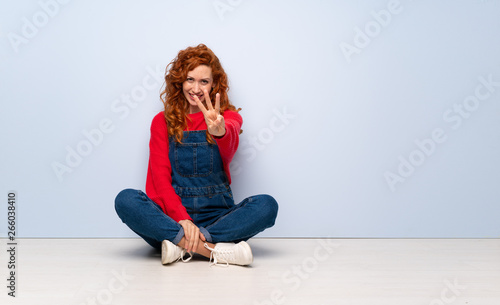 Redhead woman with overalls sitting on the floor happy and counting three with fingers