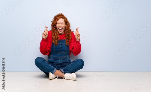 Redhead woman with overalls sitting on the floor with fingers crossing