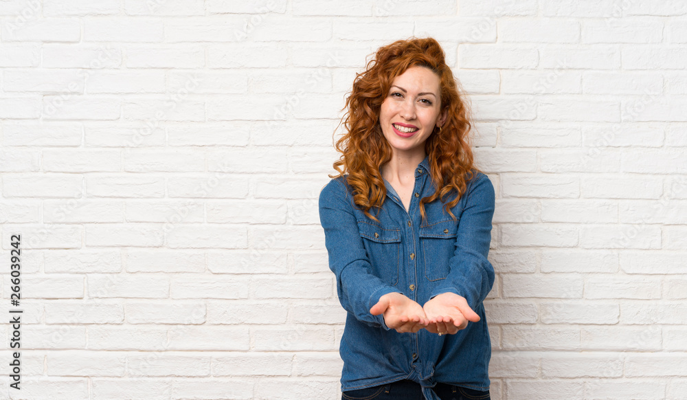 Redhead woman over white brick wall holding copyspace imaginary on the palm to insert an ad