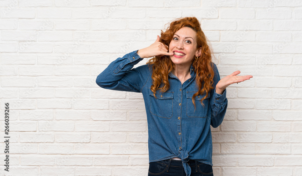 Redhead woman over white brick wall making phone gesture and doubting Redhead woman over white brick wall making phone gesture and doubting