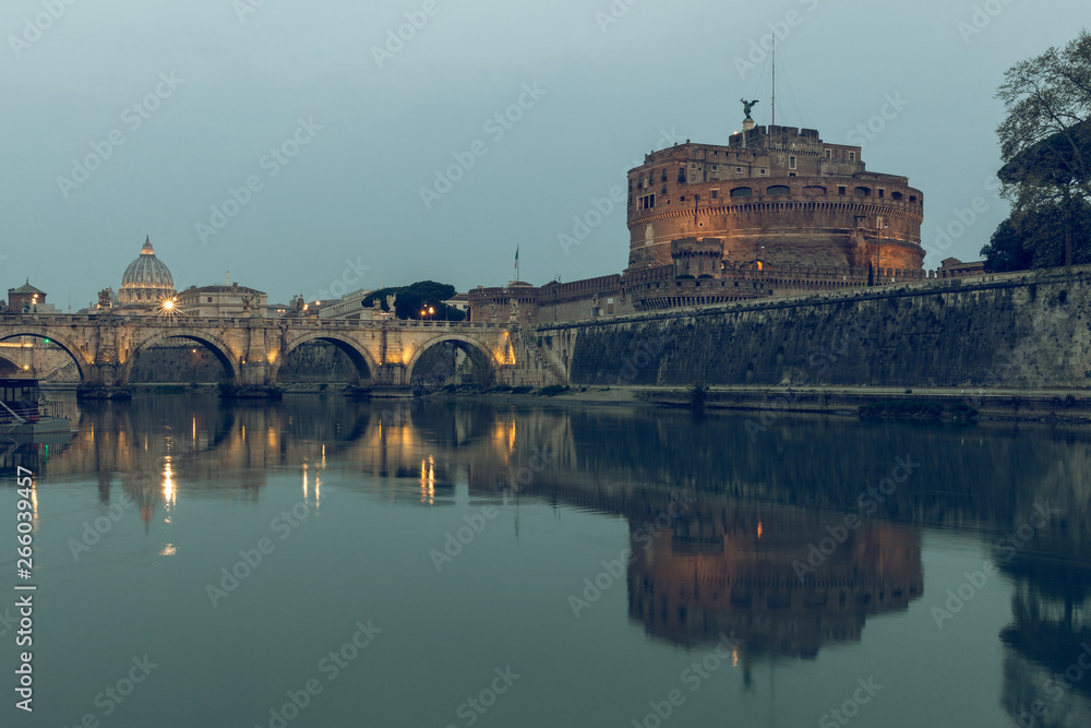 River Tiber in Rome in the background St Peters Cathedral at night. Aurelius Bridge over river with Castel Sant'Angelo at dusk. Reflections in the water of illuminated buildings and trees on the shore