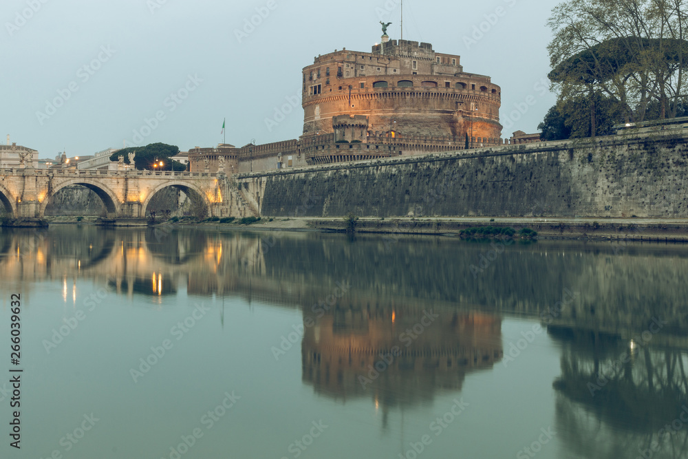 River Tiber with the Aurelius bridge over waters with Castel Sant Angelo in the morning dusk. Reflections in the water of illuminated buildings and trees on the shore
