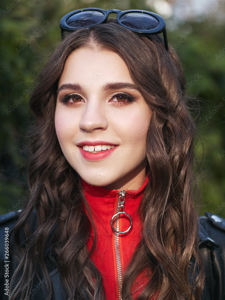 Fototapeta premium Closeup portrait of young brown-haired curly woman with trendy creative red makeup posing outdoors in bright sunlight. Pretty girl in red dress with hairstyle and makeup. 