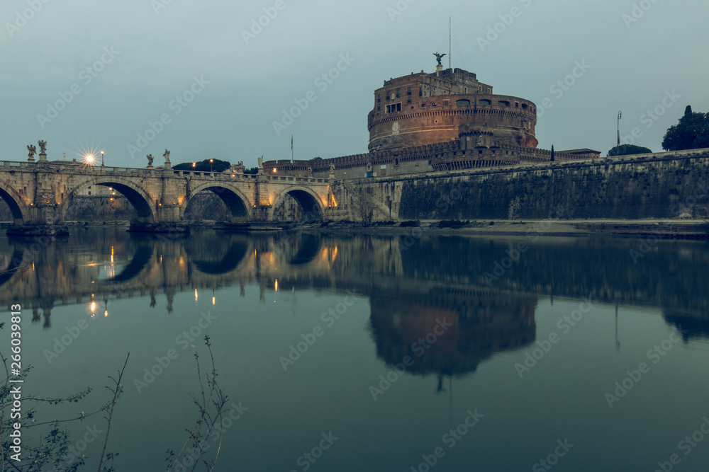 River Tiber with the Aurelius bridge over waters with Castel Sant Angelo in the morning. Reflections in the water of illuminated buildings and trees on the shore