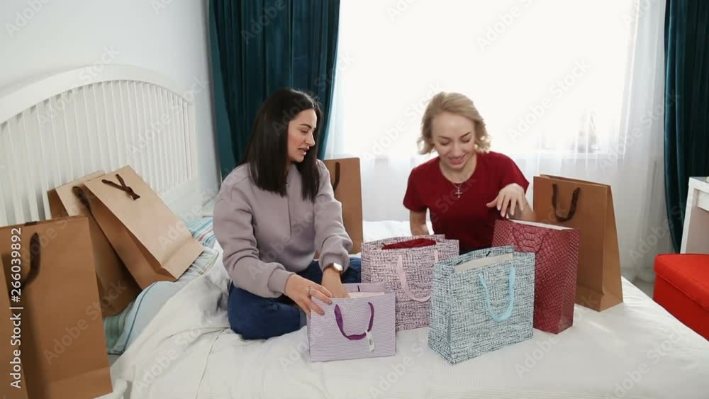 Two attractive women sit on bed and discuss new clothes from shopping. Blogging concept, recording video, friendship and consumerism concept Two attractive women sit on bed and discuss new clothes from shopping. Blogging concept, recording video, friendship and consumerism concept
