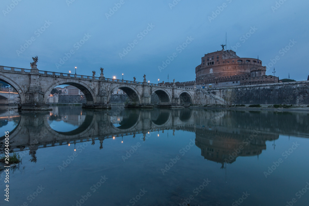 River Tiber with the Aurelius bridge over waters with Castel Sant Angelo in the morning and blue sky. Reflections in the water of illuminated buildings and trees on the shore