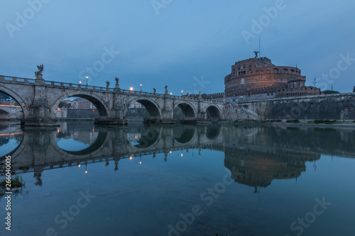 River Tiber with the Aurelius bridge over waters with Castel Sant Angelo in the morning and blue sky. Reflections in the water of illuminated buildings and trees on the shore