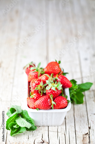 Fresh red strawberries in white bowl and mint leaves on rustic wooden background.
