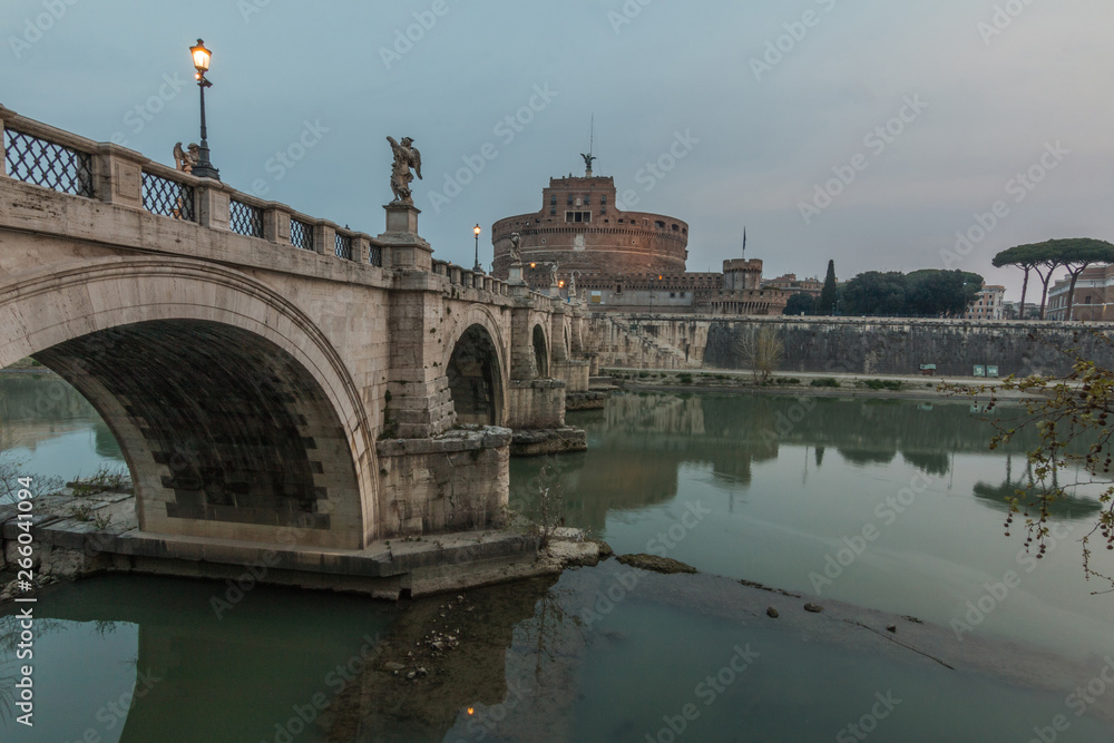 Aurelius bridge over river Tiber in the historic center of Rome with side view. Castel Sant Angelo in the background in the dusk and clouds. Historical lighting and reflections of the stone bridge in 