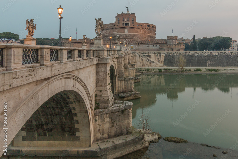 Lateral view of the Aurelius bridge with illumination over the Tiber at dawn. Stone bridge with the Castle Sant Angelo in the center of the old town of Rome. Clouds in the sky in the background