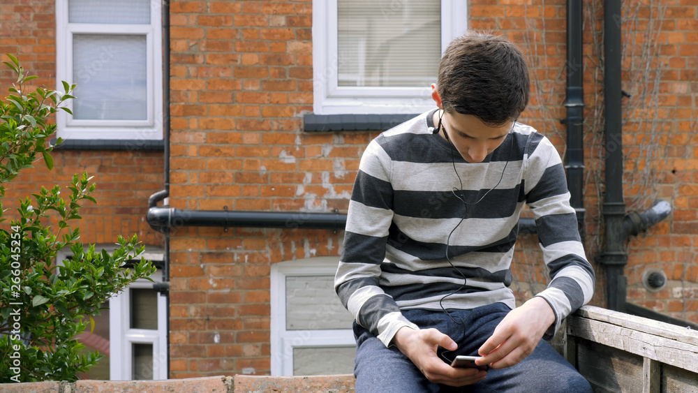 teen boy with smart phone listening or talking while sitting in british backyard garden. teenager and social media concept