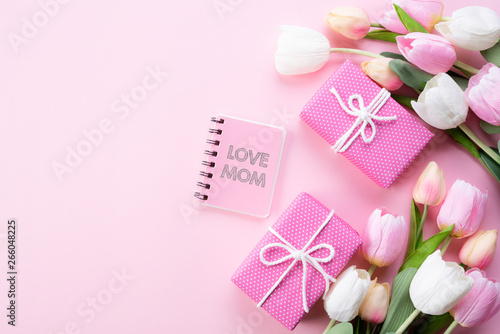 Happy mothers day concept. Top view of pink tulip flowers, gift box and note book with LOVE MOM text on pink pastel background. Flat lay.