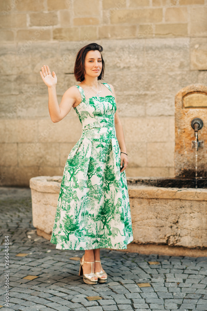 Outdoor fashion portrait of beautiful woman with dark hair, wearing long vintage styled green dress, posing on the city street