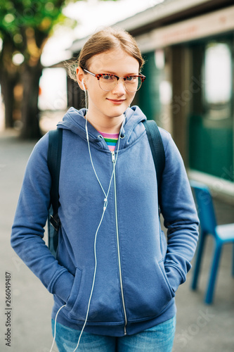 Outdoor portrait of young preteen girl wearing backpack and eyeglasses, listeming music with earphones