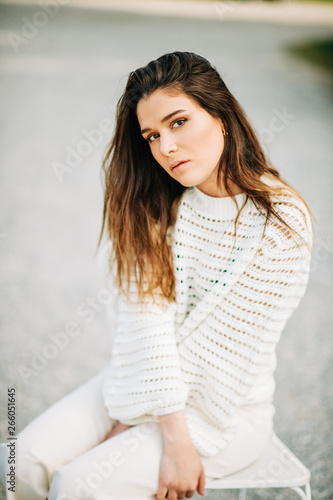 Outdoor portrait of pretty young girl wearing white pullover and jeans, sitting on the chair