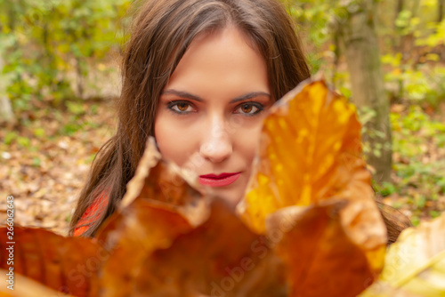 Gorgeous young woman outdoors  in a park autumn scenery  holding yellow leaves and looking focused through them at the camera. Close-up shot in natural light  retouched  vibrant colors