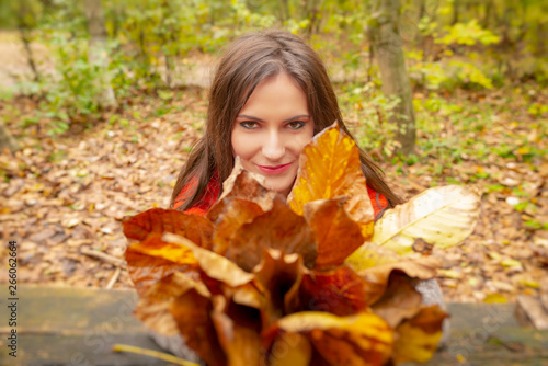 Gorgeous young woman outdoors  in a park autumn scenery  holding yellow leaves  looking at the camera and smiling. Close-up shot in natural light  retouched for perfect skin  vibrant colors