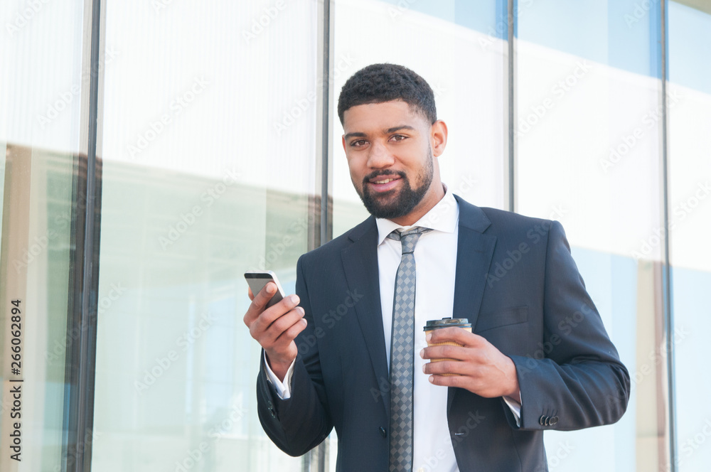 Positive handsome businessman using mobile app during coffee break outdoors. Young mix raced man in formal suit holding phone and takeaway cup and looking at camera. Smartphone using concept Positive handsome businessman using mobile app during coffee break outdoors. Young mix raced man in formal suit holding phone and takeaway cup and looking at camera. Smartphone using concept