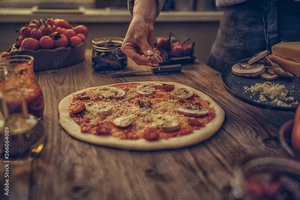 A housewife put chees on base in a rural italian kitchen. Make pizza for dinner, hands add ingriduent. Italian food making pizza with ingredients, tomatoes, chees and mushrooms on wooden tabletop.