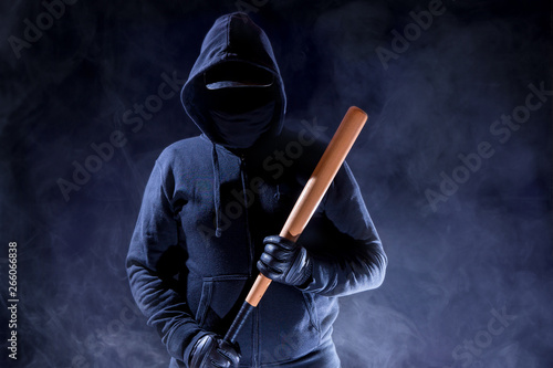 A man with a hood is standing in the dark. He is armed with a baseball bat. Concept violence.