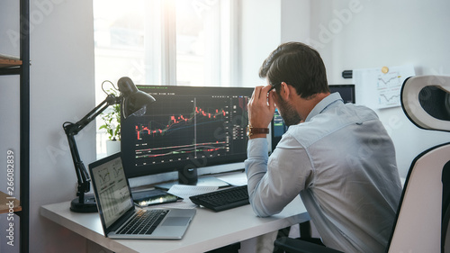 Working process. Smart male trader looking at charts on multiple computer screens and analyzing data while sitting in his modern office