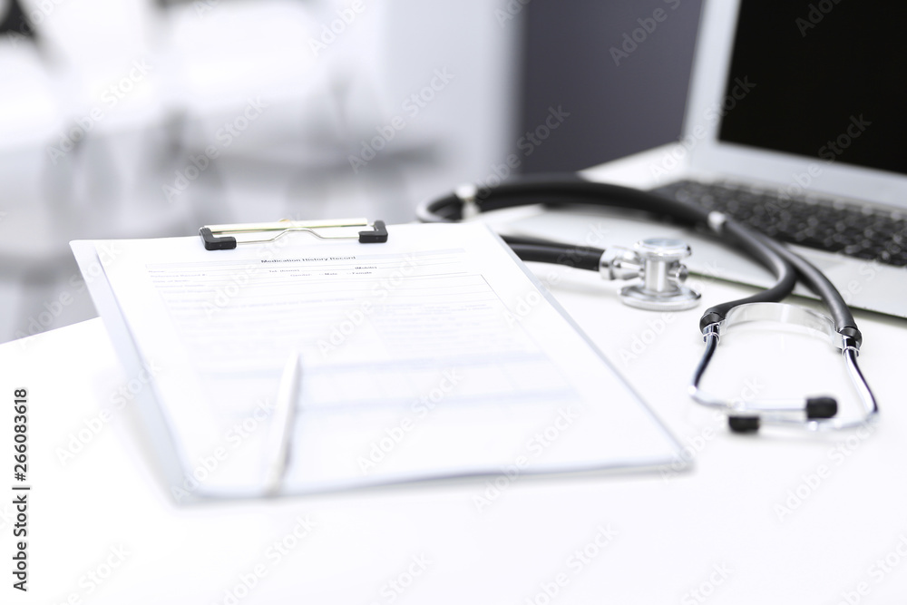 Stethoscope, clipboard with medical form lying on hospital reception desk with laptop computer and busy doctor and patient communicating at the background. Medical tools at doctor working table Stethoscope, clipboard with medical form lying on hospital reception desk with laptop computer and busy doctor and patient communicating at the background. Medical tools at doctor working table
