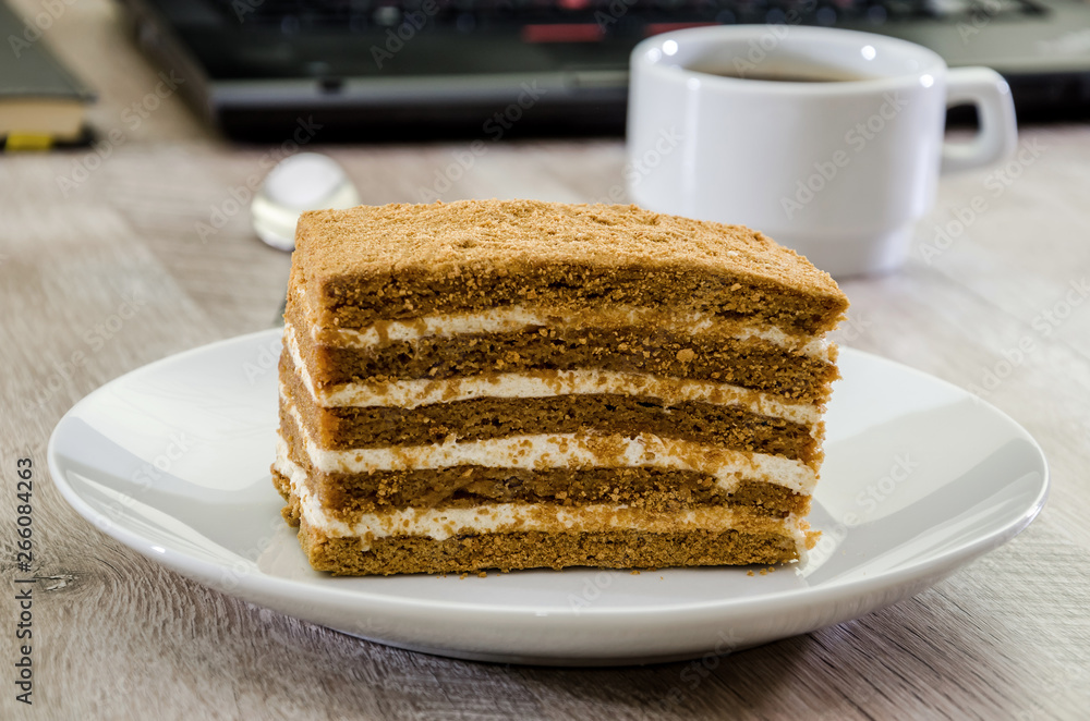 a piece of cake on a plate and a cup of coffee on the background of a laptop and notebook a piece of cake on a plate and a cup of coffee on the background of a laptop and notebook