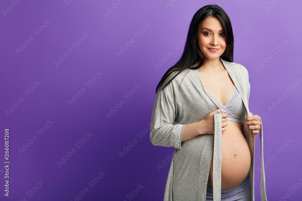 Expectant mother opens a little her dressing gown to expose her round belly, looking at camera with beaming smile, enjoys good moment of pregnancy Expectant mother opens a little her dressing gown to expose her round belly, looking at camera with beaming smile, enjoys good moment of pregnancy