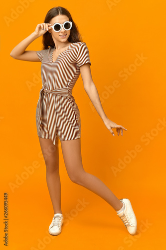 Vertical shot of stylish fashionable young woman with tanned skin, long slim legs posing in orange studio wearing white sneakers, striped summer jumpsuit and stylish sunglasses, having happy look