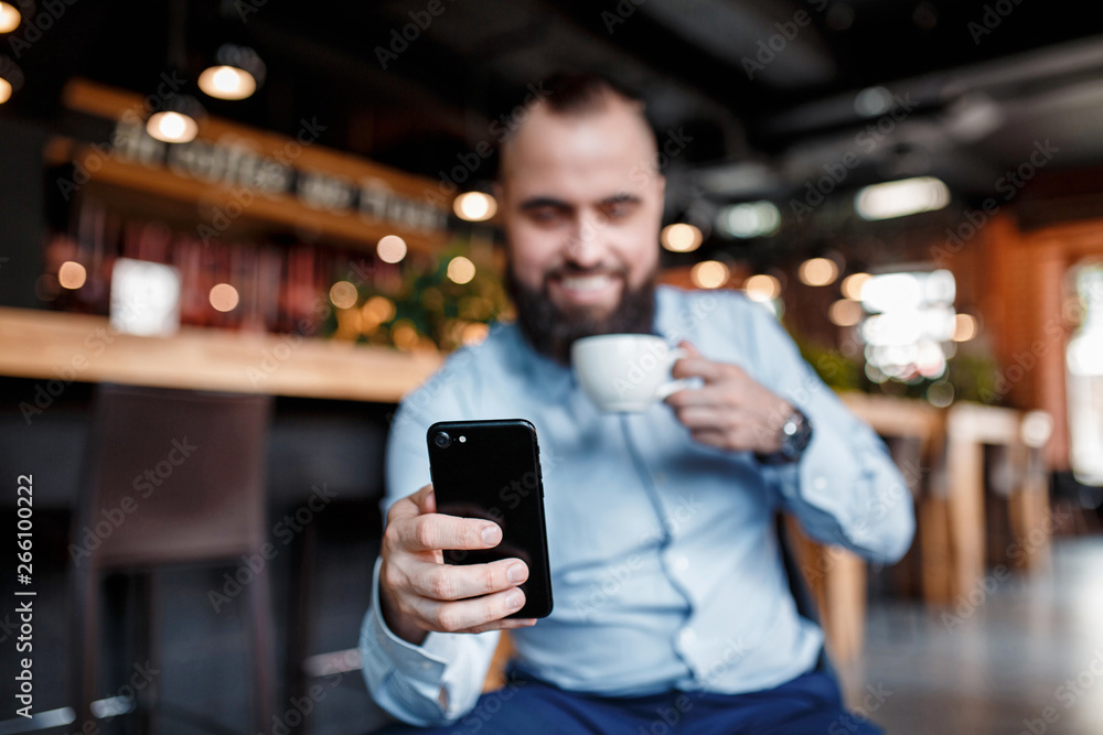 In the morning, the businessman follows news, the stock price via the telephone and drinks coffee. young investor. Bearded man with a smile looks into the phone with coffee in hand In the morning, the businessman follows news, the stock price via the telephone and drinks coffee. young investor. Bearded man with a smile looks into the phone with coffee in hand