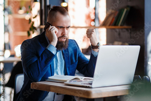 Serious bearded businessman conducts telephone conversations on a mobile phone at the computer. Unhappy man scolds on the phone. Emitations of rage in the office at the manager