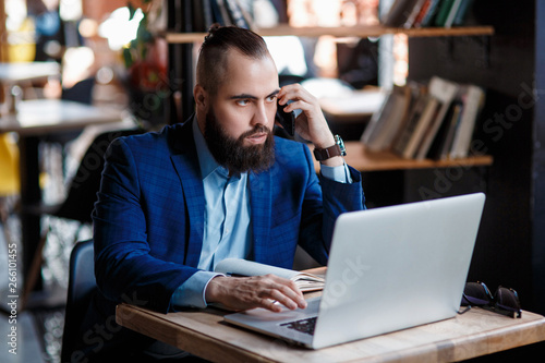 young bearded businessman at work in the office. A man sits at a laptop talking through a mobile phone. telephone conversations in coworking