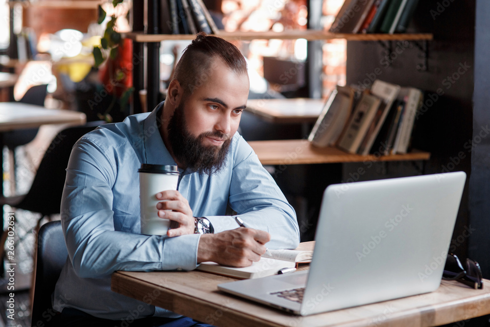 Young serious bearded businessman working on computer at table,drinking coffee.Man analyzes information, data, develops business plan. Freelancer, entrepreneur.Online marketing, education, e-learning Young serious bearded businessman working on computer at table,drinking coffee.Man analyzes information, data, develops business plan. Freelancer, entrepreneur.Online marketing, education, e-learning