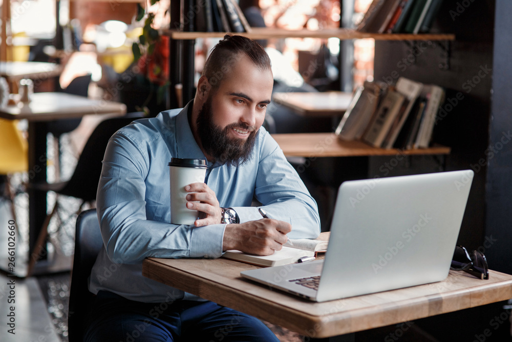 Young serious bearded businessman working on computer at table,drinking coffee.Man analyzes information, data, develops business plan. Freelancer, entrepreneur.Online marketing, education, e-learning