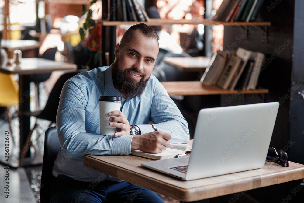 Young serious bearded businessman working on computer at table,drinking coffee.Man analyzes information, data, develops business plan. Freelancer, entrepreneur.Online marketing, education, e-learning Young serious bearded businessman working on computer at table,drinking coffee.Man analyzes information, data, develops business plan. Freelancer, entrepreneur.Online marketing, education, e-learning