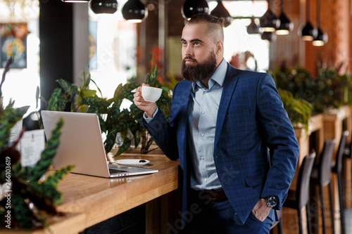 Young serious bearded businessman working on computer at table,drinking coffee.Man analyzes information, data, develops business plan. Freelancer, entrepreneur.Online marketing, education, e-learning