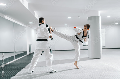 Young Caucasian boy in dobok kicking barefoot while trainer holding kick target. Taekwondo training concept.