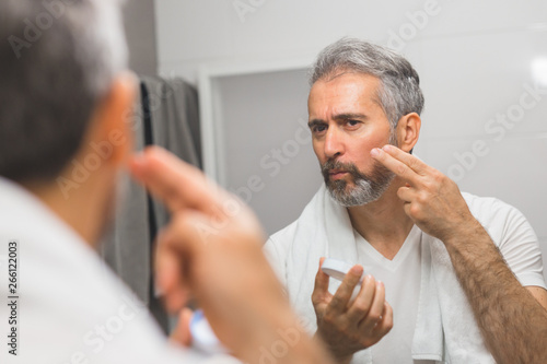 middle aged bearded gray haired man applying cream in bathroom